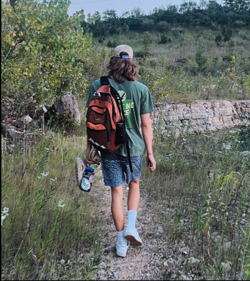 Boy with long hair wearing Vistathread Green T-shirt that says leave no trace. Has backpack on and is walking through greenery. 